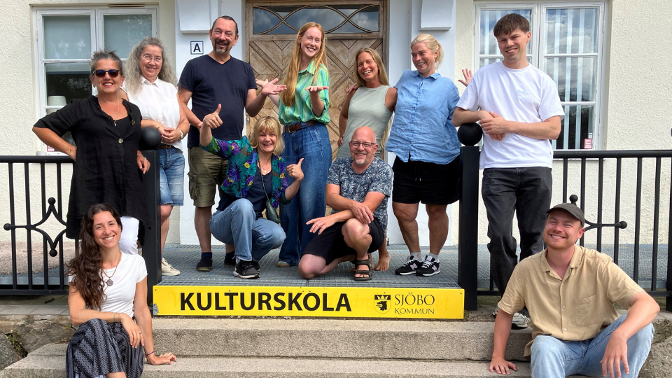 En gruppbild där 11 leende personer står och sitter på en trappa med en gul skylt där det står "kulturskolan"
