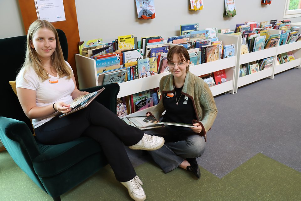 Linn sitter i en fåtölj och Maria sitter på golvet med benen i kors. Båda har barnböcker i knäet. De är på biblioteket i Sjöbo.