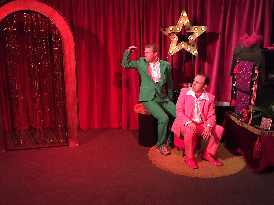 Två skådespelare, en i grön kostym och en i rosa kostym, sitter i ett rött scenrum och spanar efter någonting