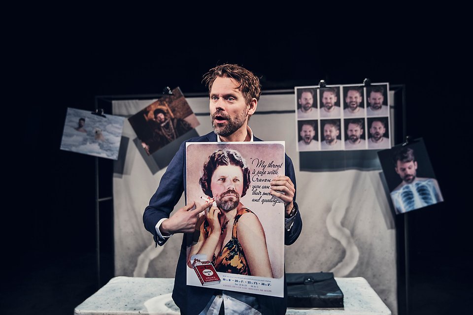 En skådespelare håller upp en affisch med sitt eget ansikte photoshopadet på en annan människas kropp. I bakgrunden syns en massa bilder uppsatta på en vägg. 