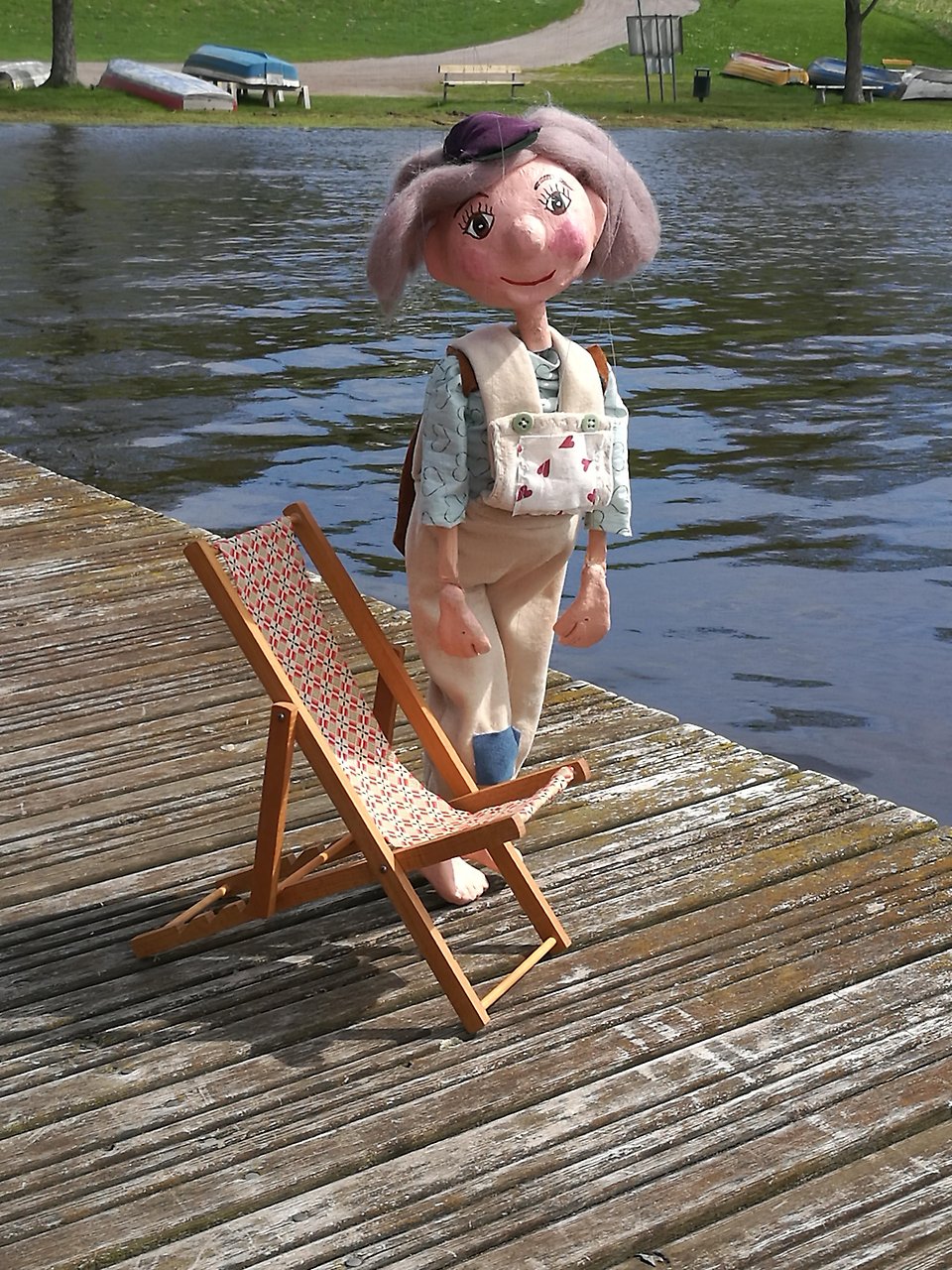 Ett foto på en glad marionettdocka med grått hår och beiga snickarbyxor som står på en brygga. I bakgrunden ser man vattnet länge bort en strand