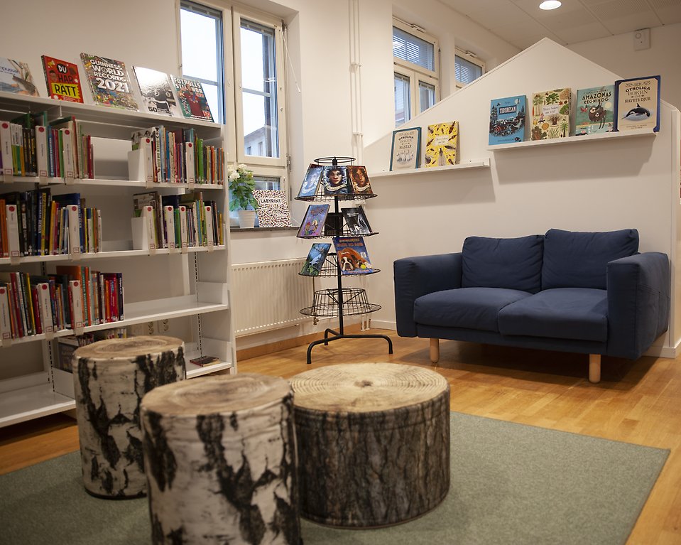 Interiör från Blentarps bibliotek. En soffa och puffar som ser ut som trästubbar. Barnböcker i hyllor vid sidan.
