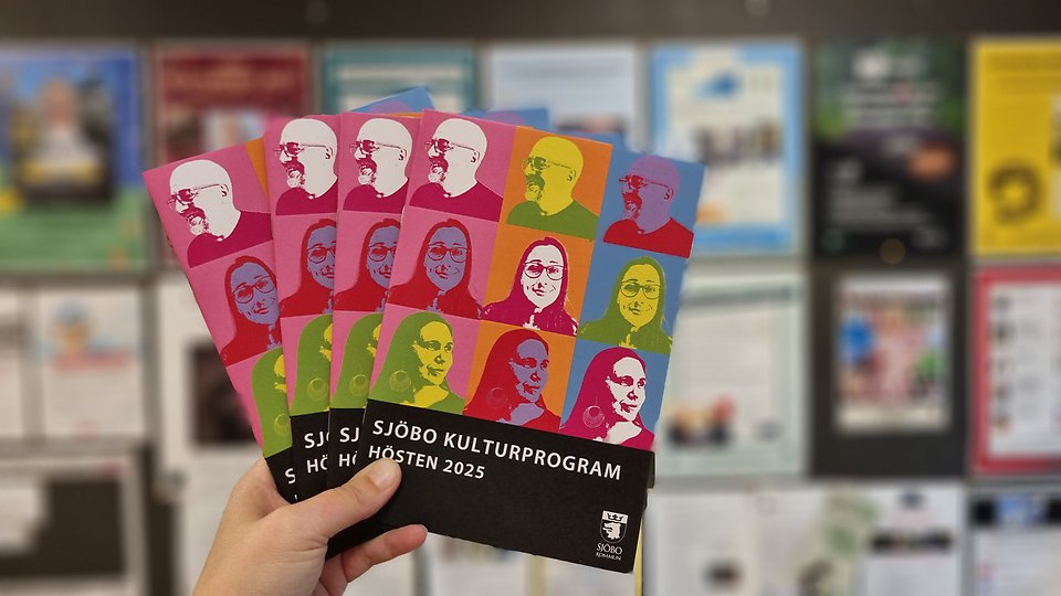 En hand håller upp fler exemplar av kulturprogrammet, i bakgrunden syns suddiga affischer