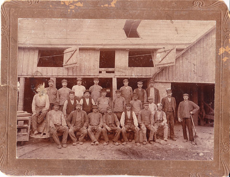 Arbetarna på tegelbruket uppställda inne på området för en gruppbild. Ett sepiafärgat fotografi.
